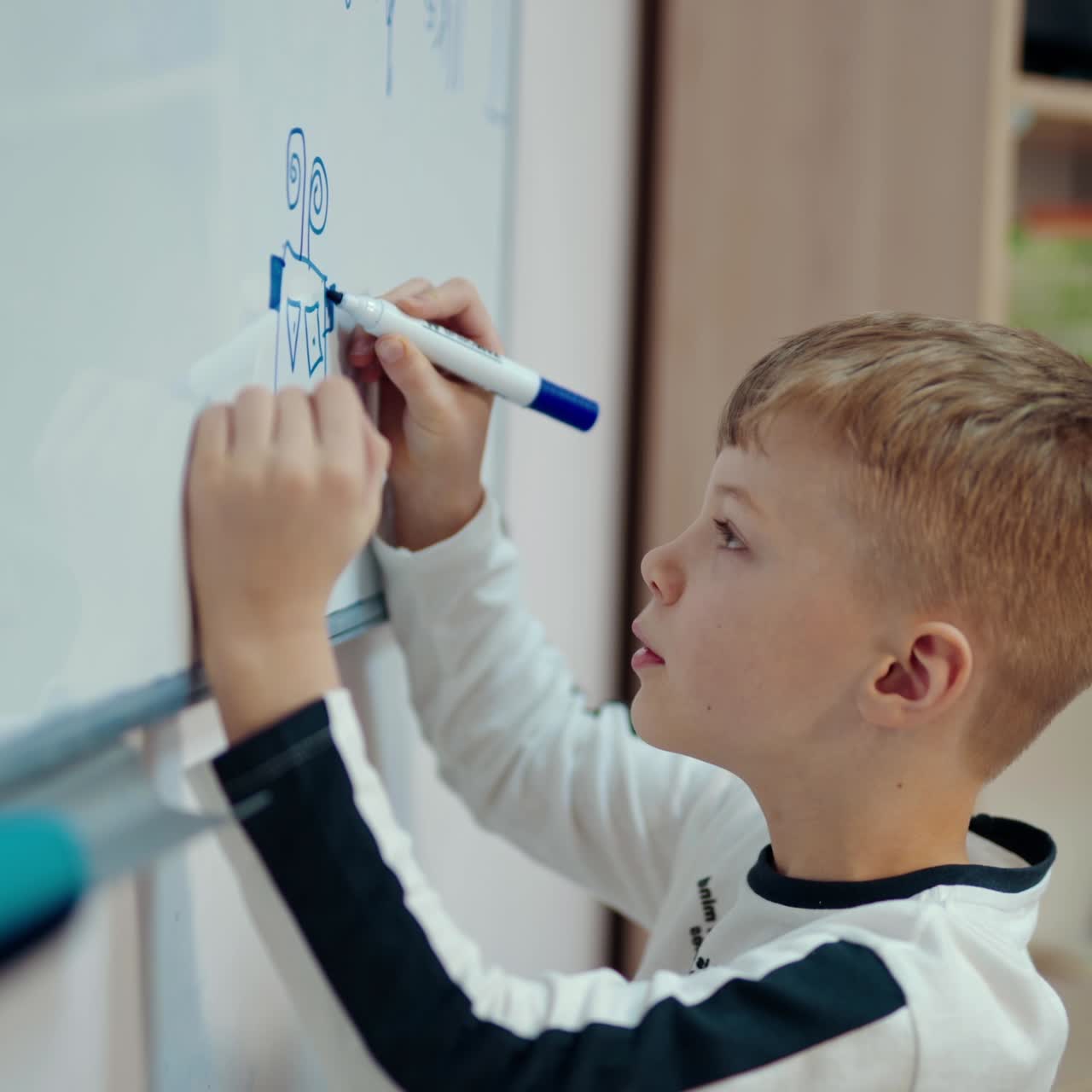 Schoolboy draws on white blackboard. Side view of a little boy writing with a marker on magnetic board in the classroom. Elementary education.