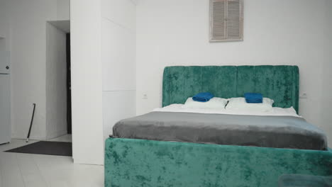 view from door of modern hotel room with green velvet bed, fresh towels on bed, and white walls. kitchen area visible in background, creating cozy and inviting atmosphere
