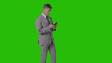 Three-Quarter-Length-Studio-Shot-Of-Businessman-In-Suit-Looking-At-Mobile-Phone-Against-Green-Screen-