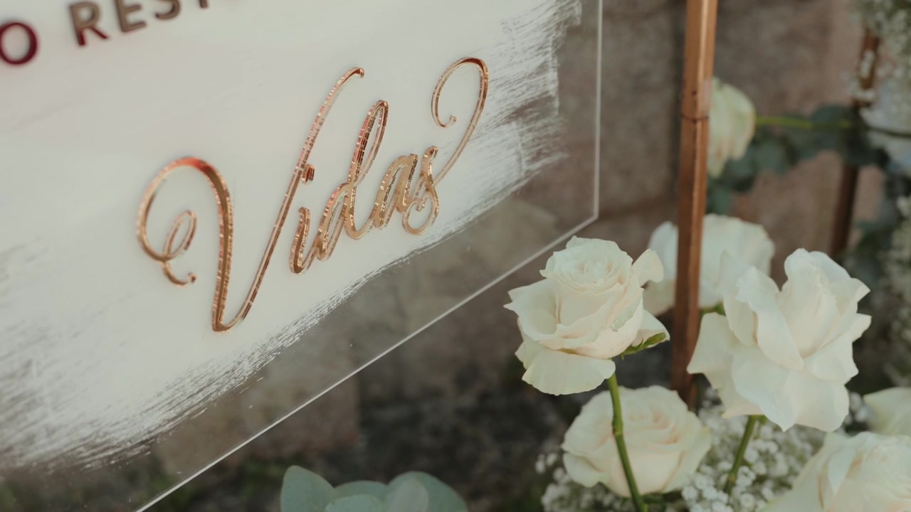 Calligrafia color oro rosa su un cartello nuziale in acrilico trasparente accanto a rose bianche e gypsophila.
