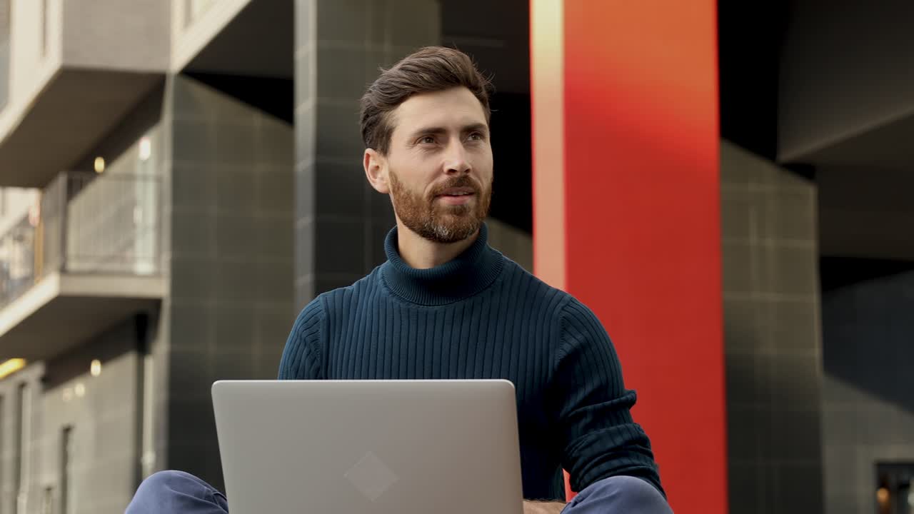 hombre joven guapo en suéter azul sentado cerca del centro de la oficina y trabajando en la computadora portátil. feliz hombre ocupado pasando el tiempo de ocio para trabajar al aire libre.