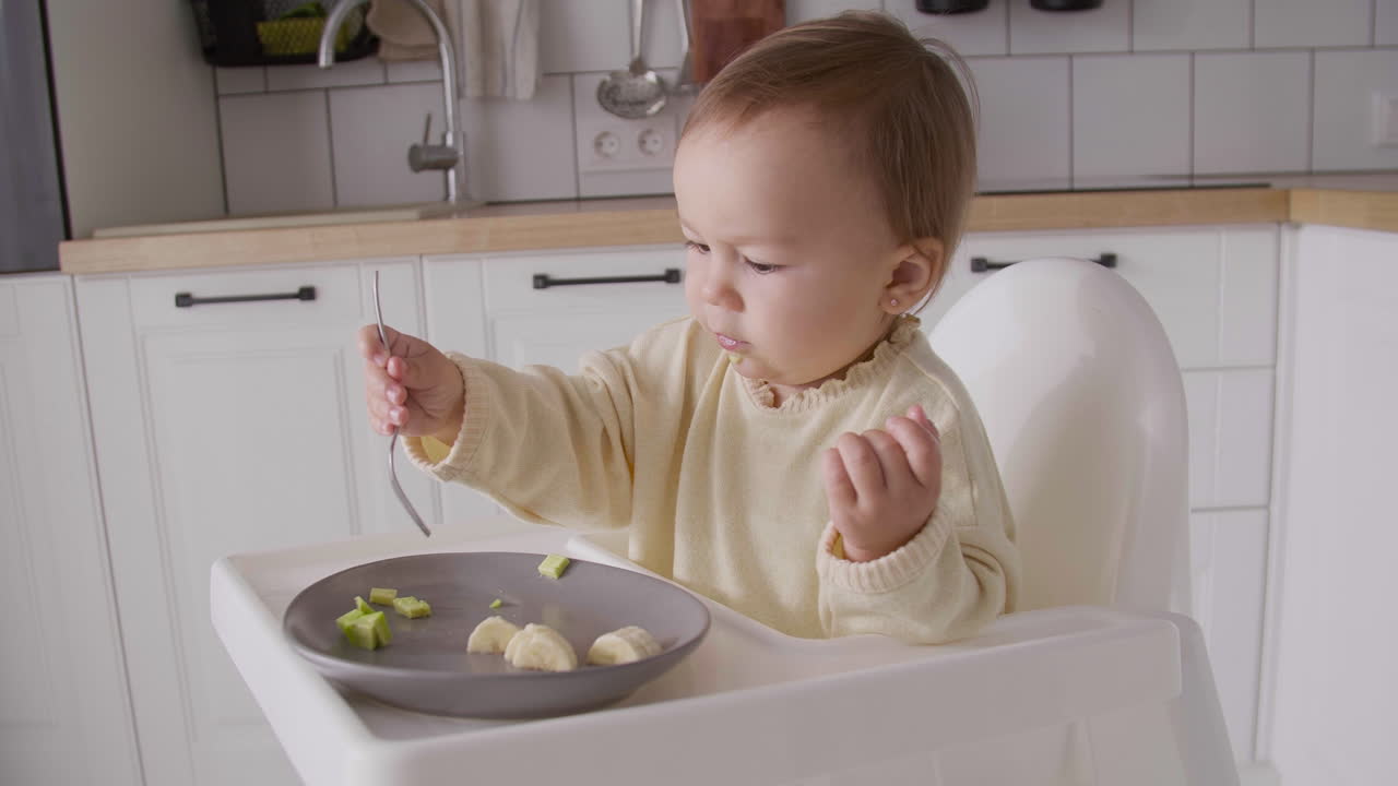 Cute Baby Girl Eating Avocado Slices Using Fork While Sitting In Her High Chair In The Kitchen