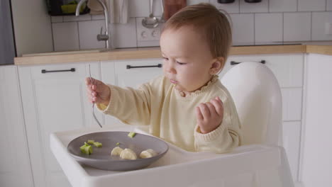 cute baby girl eating avocado slices using fork while sitting in her high chair in the kitchen