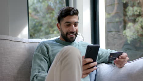 Biracial-man-sitting-on-sofa-using-smartphone-for-online-shopping-at-home,-slow-motion