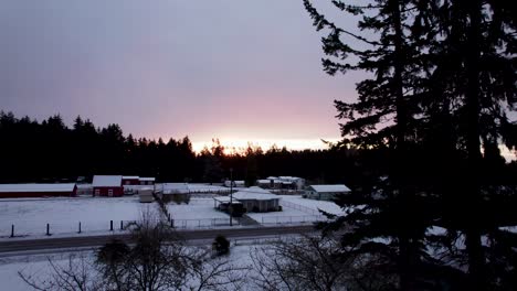 Orange-sunlight-pierces-through-snow-covered-trees,-dark-gray-skies,-suburban-area,-aerial-reveal