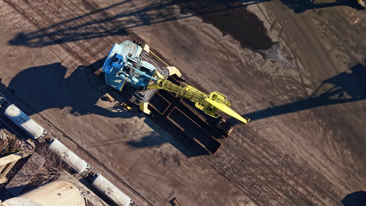 Grande macchina opera in un sito industriale durante il giorno, spostando materiali a terra vicino a container di stoccaggio.