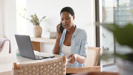 Feliz-Mujer-Afroamericana-Sentada-En-La-Mesa-Hablando-Por-Teléfono-Inteligente-Y-Usando-Una-Computadora-Portátil,-Cámara-Lenta