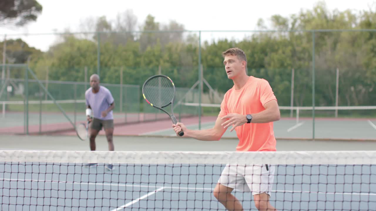 두 명의 다양한 남성 친구가 더블을 치며 느린 움직임으로 야외 코트에서 공을 네트 위로 돌려줍니다.