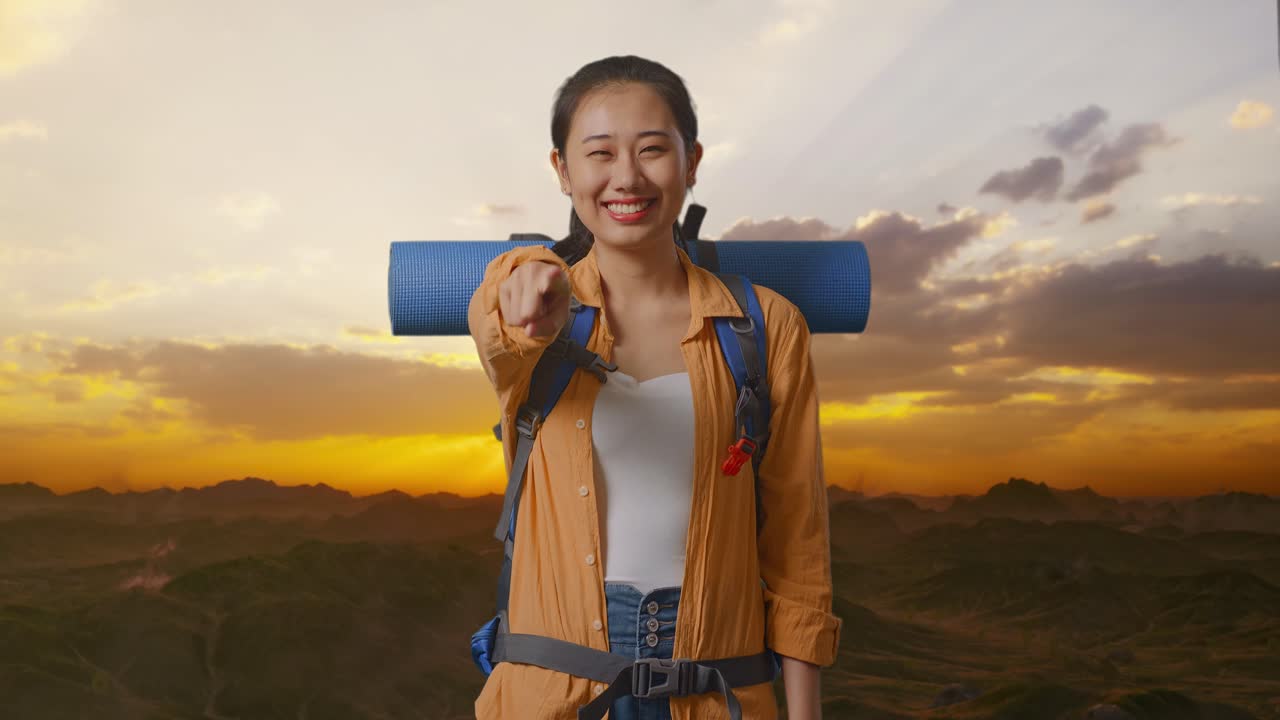 Asian Female Hiker With Mountaineering Backpack Smiling, Touching Her Chest, And Pointing To Camera While Standing On The Top Of Mountain During Sunset Time