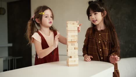 Two-preteen-girls-friends-children-playing-board-game-Jenga-on-table-in-kitchen-at-home,-slow-motion