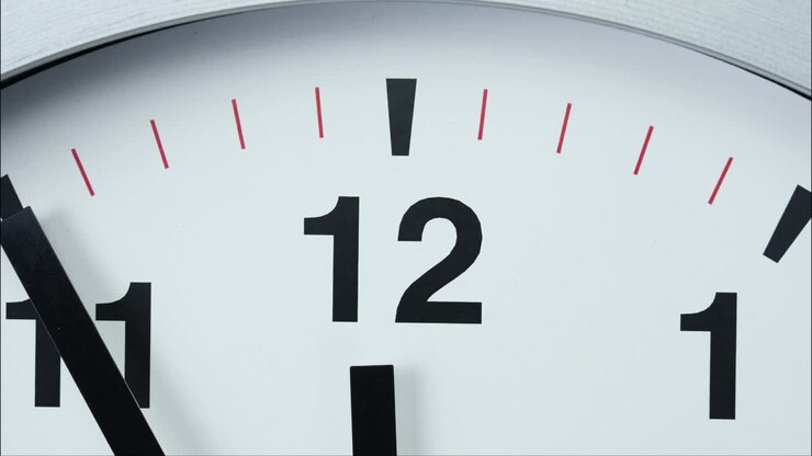 Close-up white wall clock short hand points to the number 12.