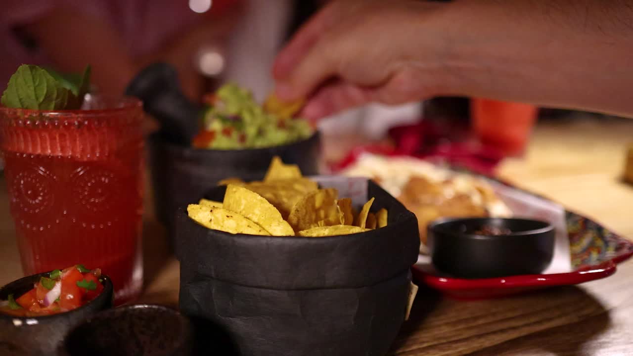 A hand reaches for nacho chips, dips them in guacamole, and enjoys fresh salsa beside a red cocktail in a warmly lit restaurant setting
