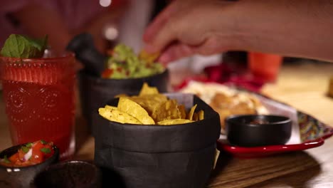 a hand reaches for nacho chips, dips them in guacamole, and enjoys fresh salsa beside a red cocktail in a warmly lit restaurant setting
