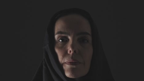 a muslim woman with a somber expression walks from the darkness