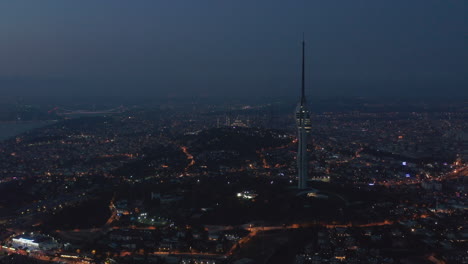 Amplia-Vista-De-La-Enorme-Nueva-Torre-De-Televisión-De-Estambul-Con-Mezquita-En-La-Colina-En-El-Fondo-Por-La-Noche,-Toma-Aérea-De-Drones