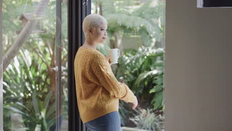 Happy-biracial-woman-drinking-coffee-at-window-at-home-in-slow-motion