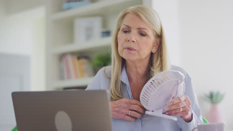 mujer madura en la menopausia que tiene aleteos de calor en casa enfriándose con un ventilador conectado a una computadora portátil