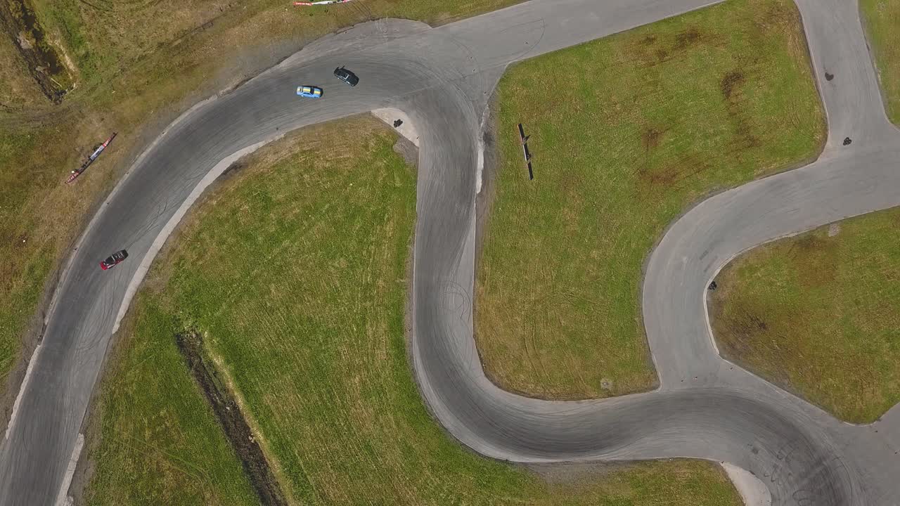Stillbillede fra luften, set direkte ovenfra, af en racerbane med driftingbiler, rygende dæk og solrigt vejr.
