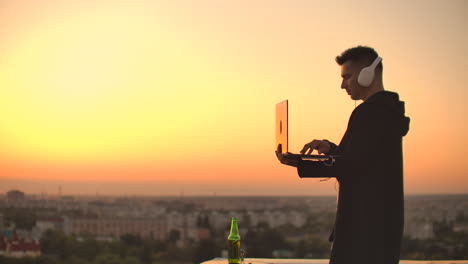 usando un portátil en el techo con vista a la ciudad y gráfico de divisas. concepto de hacking y estadísticas. un hombre al atardecer en cámara lenta escribiendo código de software en un portátil hacker