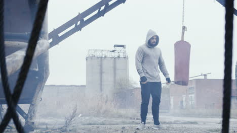 hombre caucásico con capucha gris y terminando su entrenamiento de boxeo cerca del saco de boxeo rojo al aire libre en una fábrica abandonada en una mañana nublada