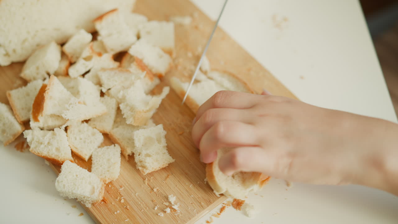 High angle close up of female hands chopping bread slices into precise small pieces on wooden board over white table under soft window light, crumbs scattered, focus on calm culinary preparation