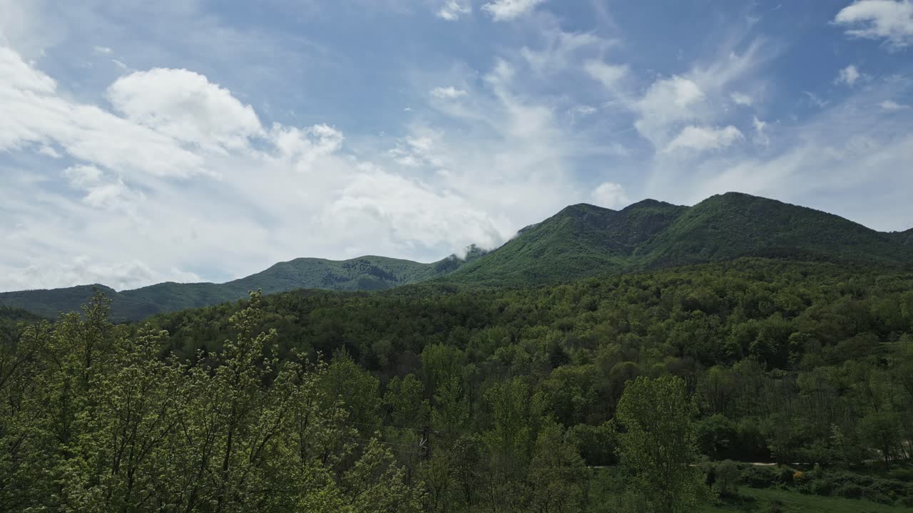 Verdant mountain range stretching beneath drifting white clouds, casting soft shadows across lush green valleys bathed in bright sunlight and serene natural beauty