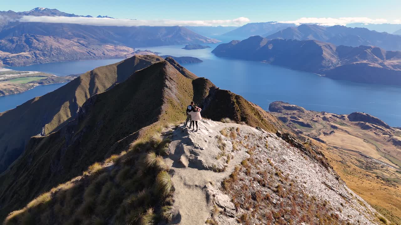 4K drone captures a couple walking up to a famous viewpoint at Roy’s Peak in Wanaka, New Zealand. The couple take in the stunning lake and mountain views all around