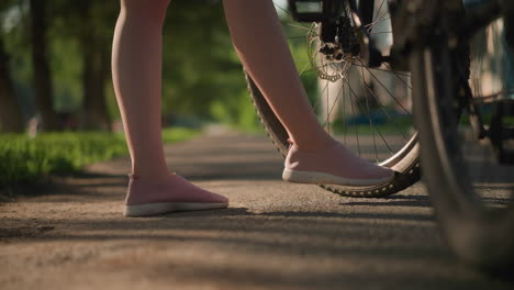 side view of individual legs in pink sneakers kicking bicycle tire to check air pressure, lush greenery and trees create beautiful backdrop, with shadow details and car passing in blurred background