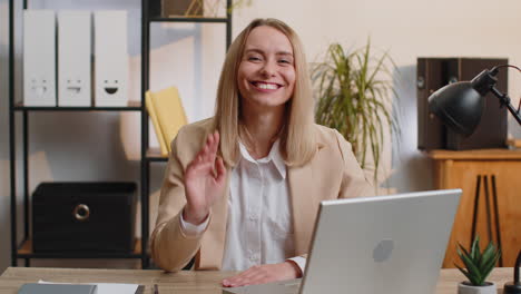 businesswoman works on laptop smiling friendly at camera and waving hands gesturing hello at office