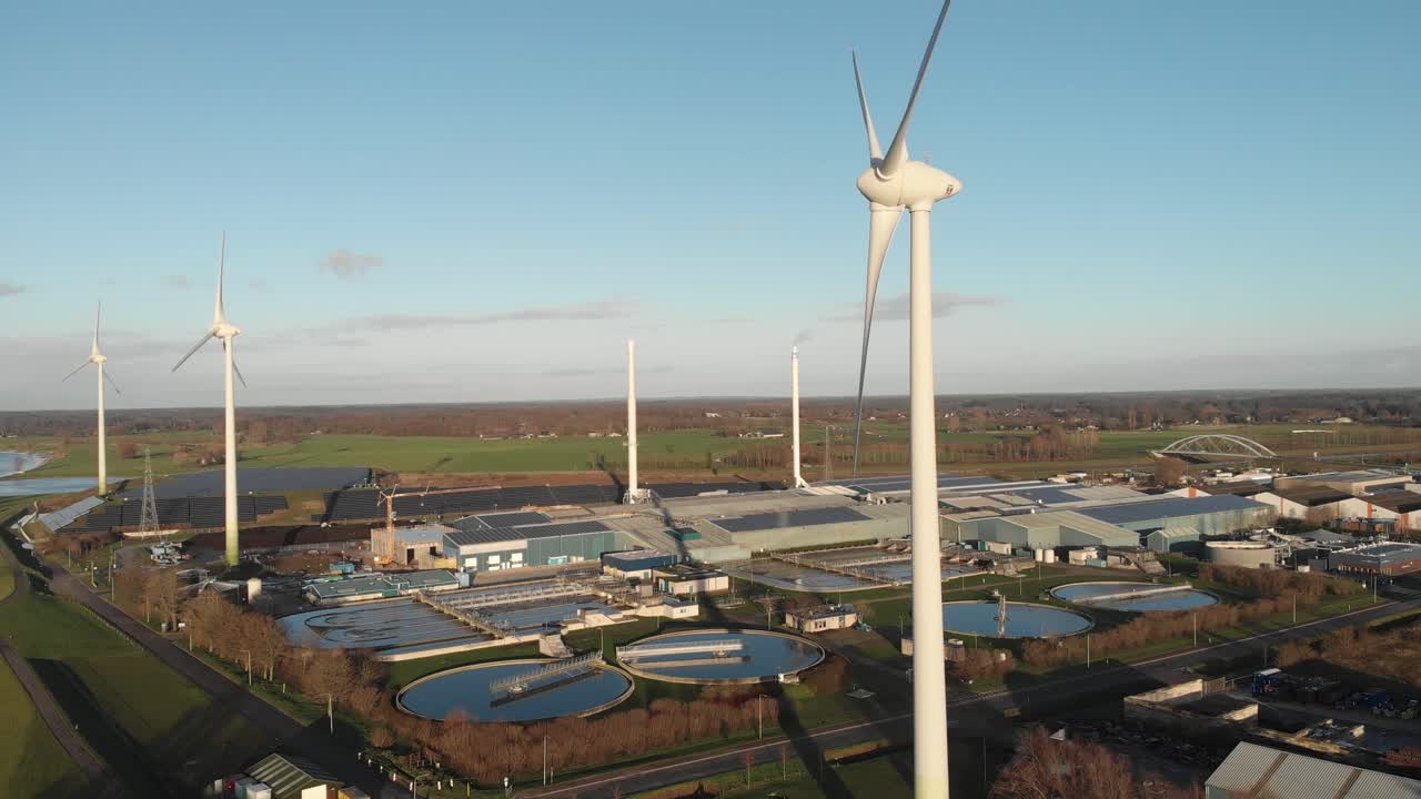 Spinning wind turbines generating sustainable energy with a water treatment facility, bio energy and fields of solar panels in industrial area in the background at a river delta landscape at sunset