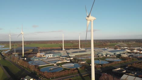 spinning wind turbines generating sustainable energy with a water treatment facility, bio energy and fields of solar panels in industrial area in the background at a river delta landscape at sunset