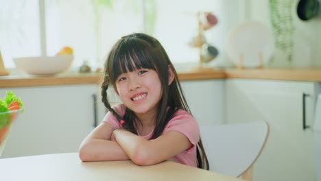 Retrato-De-Un-Niño-Pequeño-Asiático-Sonriendo-Y-Mirando-A-La-Cámara-En-La-Cocina.-Las-Niñas-Encantadoras-Se-Sientan-En-La-Mesa,-Se-Sienten-Felices-Y-Disfrutan-Del-Fin-De-Semana-Matutino-En-Casa.-Concepto-De-Estilo-De-Vida-De-Actividad-De-Ocio-En-El-Hogar.