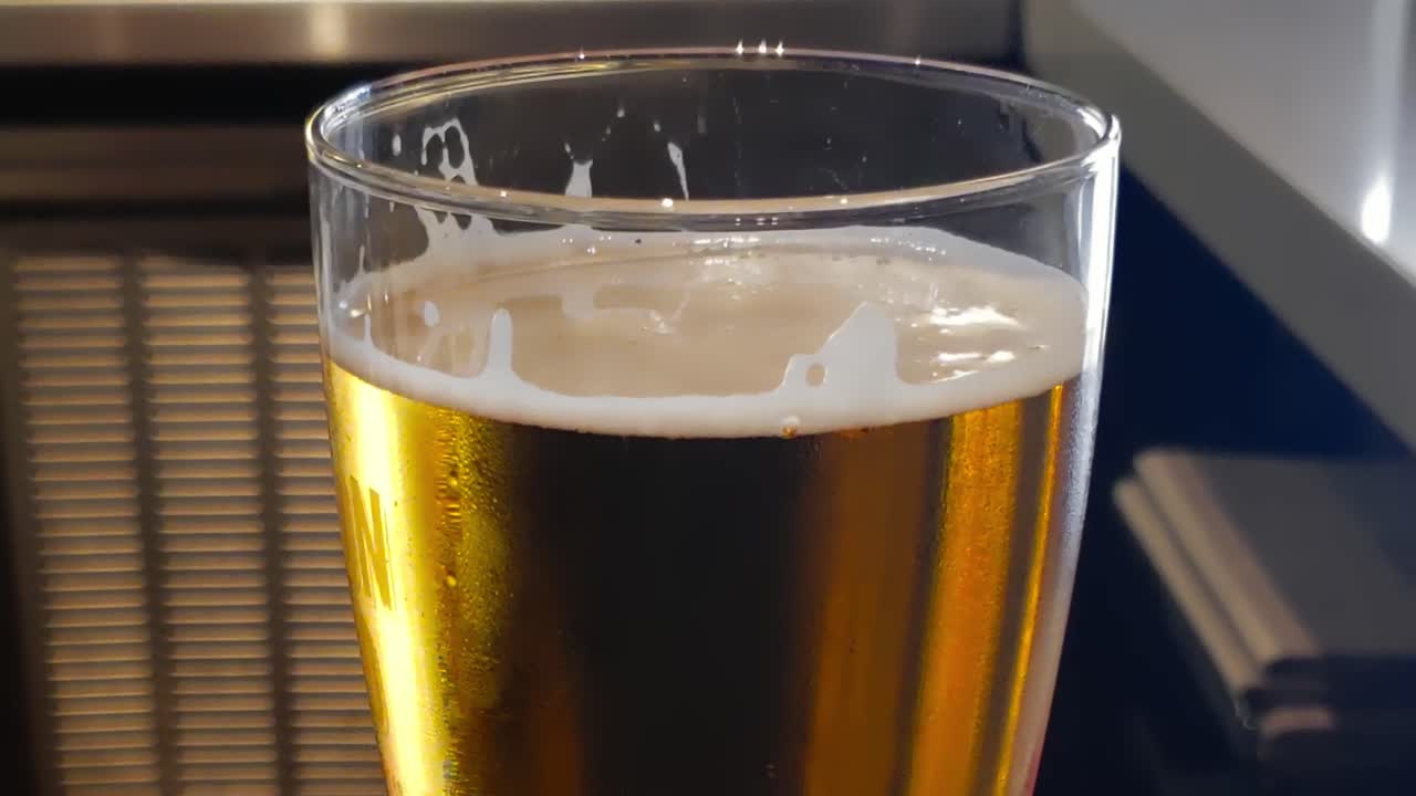 cierre en cámara lenta de la parte superior de un vaso de pinta de cerveza lager naranja dorada en una esquina de barra de mármol brillante reflectante y un fondo de una mini nevera de suministro de bebidas