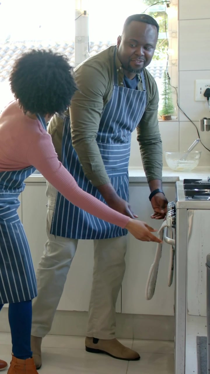 Vertical video: Leaning pair in striped aprons adjusting oven control at kitchen range with whisk