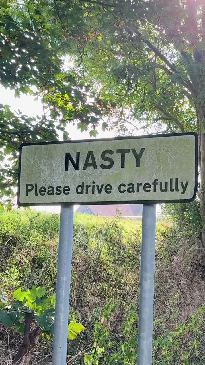 Vertical slow-motion pan of Nasty, Hertfordshire road sign. “Please drive carefully” is visible underneath, offering a literal traffic warning and a playful metaphorical concept for risk and caution