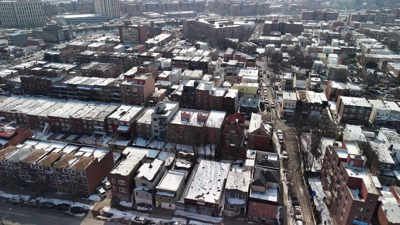 Aerial footage moving over a dense urban residential neighborhood with snow covering the rooftops and streets, showcasing apartment buildings and houses on a sunny but cold winter day