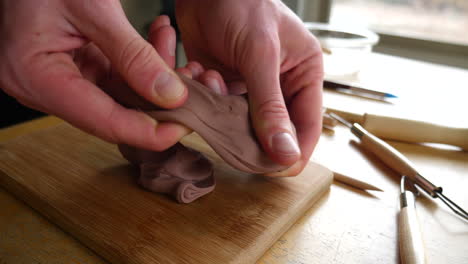 An-artist-pulling-a-piece-of-brown-modeling-clay-and-creating-a-sculpture-with-his-hands-and-tools-in-an-art-studio-classroom