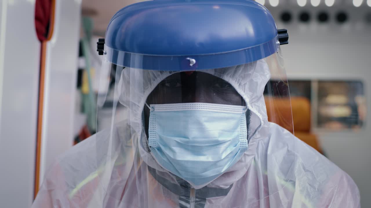 african American Paramedic in Hazmat Suit Looking at Camera Weary Black Male Medical Practitioner in Hazmat Suit with Mask and Face Shield Looking at Camera while Riding Ambulance to Emergency during Pandemic