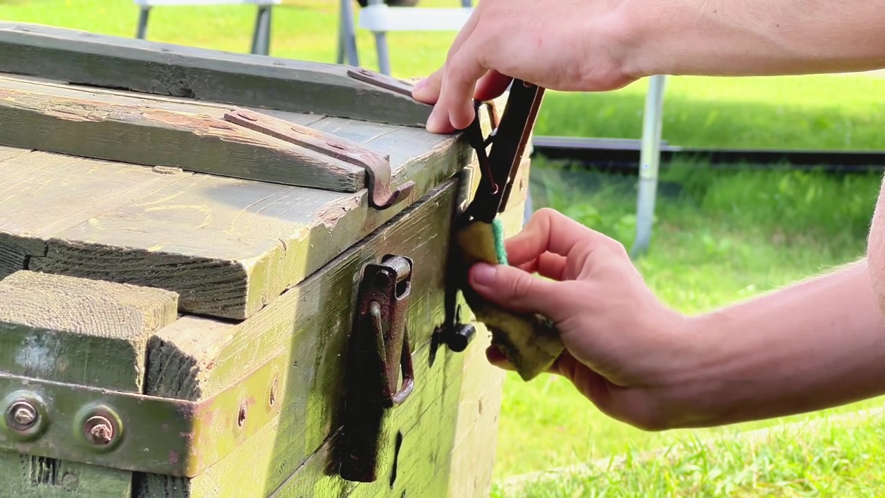 Close-up shot of hands wiping down a vintage green wooden ammo crate outdoors. Ideal for restoration tutorials, DIY projects, historical documentary content, and craft videos.