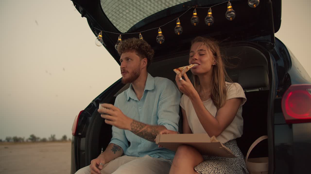 A bearded man with curly hair in a blue shirt is drinking tea next to his blonde girlfriend, who is eating pizza, they are sitting in the trunk of the black car decorated with the lights against the background of the flying birds in the yellow sky