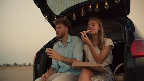 A-bearded-man-with-curly-hair-in-a-blue-shirt-is-drinking-tea-next-to-his-blonde-girlfriend,-who-is-eating-pizza,-they-are-sitting-in-the-trunk-of-the-black-car-decorated-with-the-lights-against-the-background-of-the-flying-birds-in-the-yellow-sky