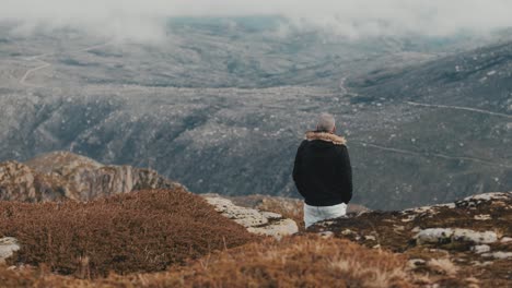 A-thoughtful,-handsome-man-with-short-hair-and-a-mustache-walks-slowly-through-the-stunning,-mountainous-landscape,-hands-in-pockets,-with-clouds-on-the-horizon