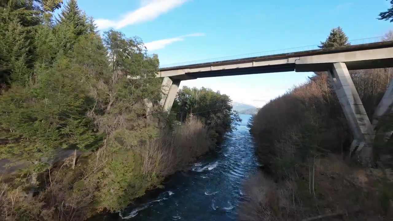 A breathtaking aerial perspective flying over the crystal-clear turquoise waters of the Correntoso River, showcasing both the old wooden pedestrian bridge and the modern concrete highway crossing.