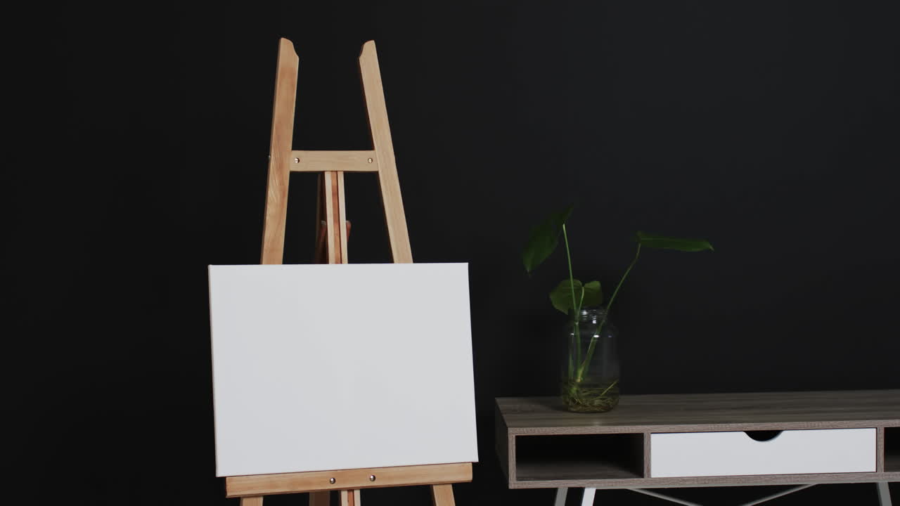 Video of white canvas sign on wooden easel with copy space and plant on desk on black background