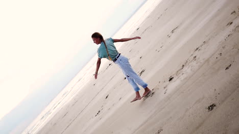 Happy-blond-girl-with-long-hair-in-a-braid,-dressed-with-sportswear,-doing-cartwheels-and-spinning-on-the-seashore