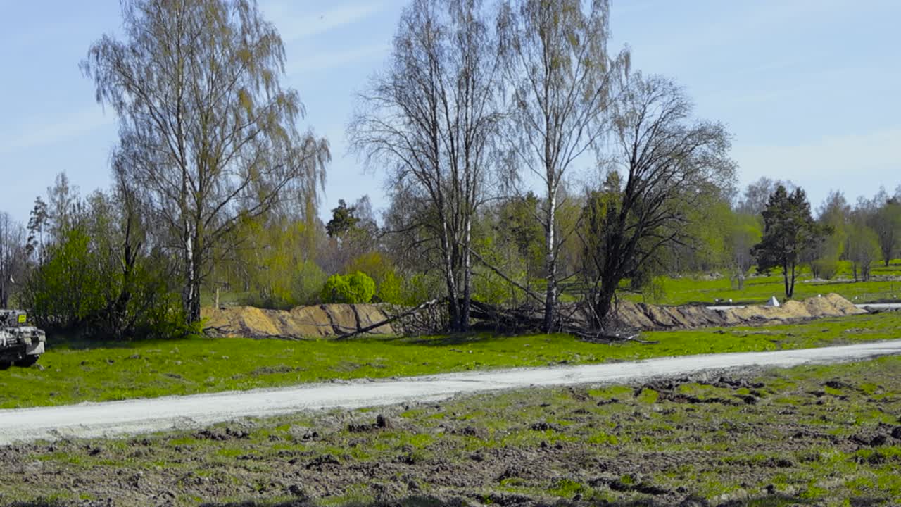 Panning footage of a Challenger 2 4034 British army armoured tank with a cannon on a grassy green field aiming at enemy lines or trenches during a sunny summer day with green trees around and in back.