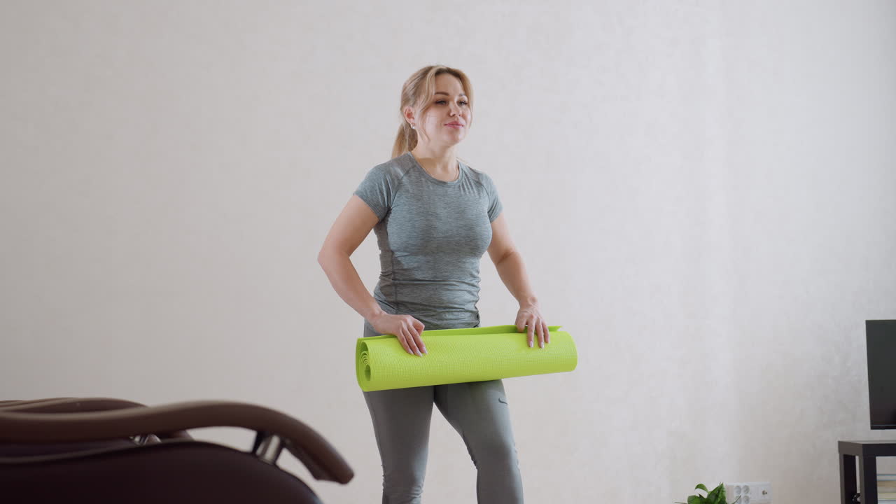 Young woman in grey workout clothes holds bright green exercise mat indoors with determined expression, preparing for home workout in minimalist room