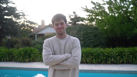 young caucasian man with arms crossed over chest, standing by the poolside and waves to camera