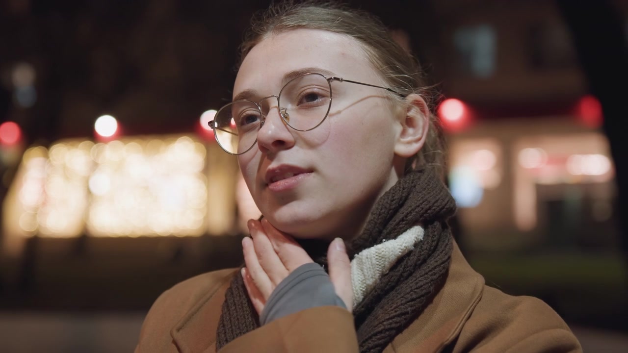 Evening street portrait of white woman smiling, adjusting scarf, glasses catching light, gentle expression, warm coat, urban bokeh, candid closeup, youthful student energy, soft glow