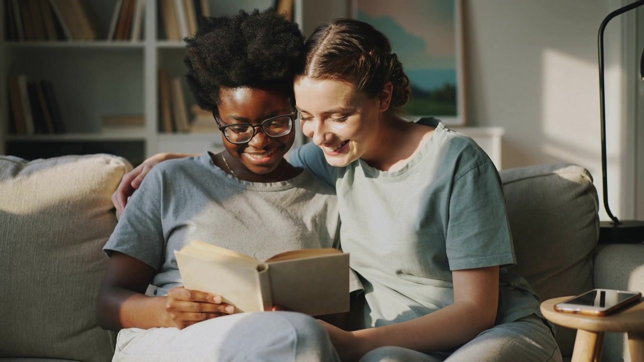 Due donne con occhiali e maglie casual, che tengono un libro tascabile aperto, appoggiate sul divano, creando un legame attraverso la lettura